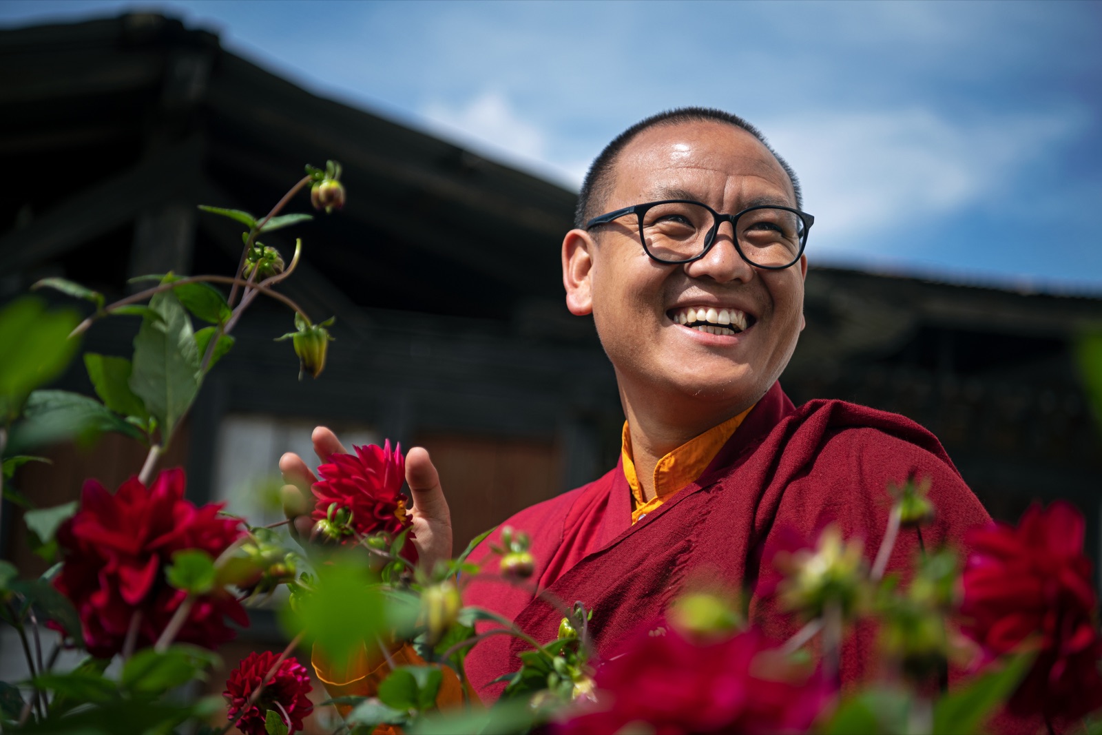 Khedrup Rinpoche, smiling, among red dahlias outside a temple in Bhutan.